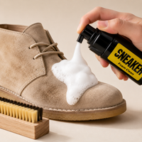 A beige suede sneaker is shown with white foam being sprayed onto it from a black bottle labeled 'SNEAKER Foam Clean'. A hand holds the spray bottle, and a wooden cleaning brush is visible in the lower left on a warm beige background.